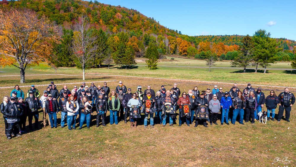 Members of American Legion Riders Post 259 and Red Knights 44 gathered together in a field with bright fall foliage covering the surrounding hills, many wearing leather vests with club patches