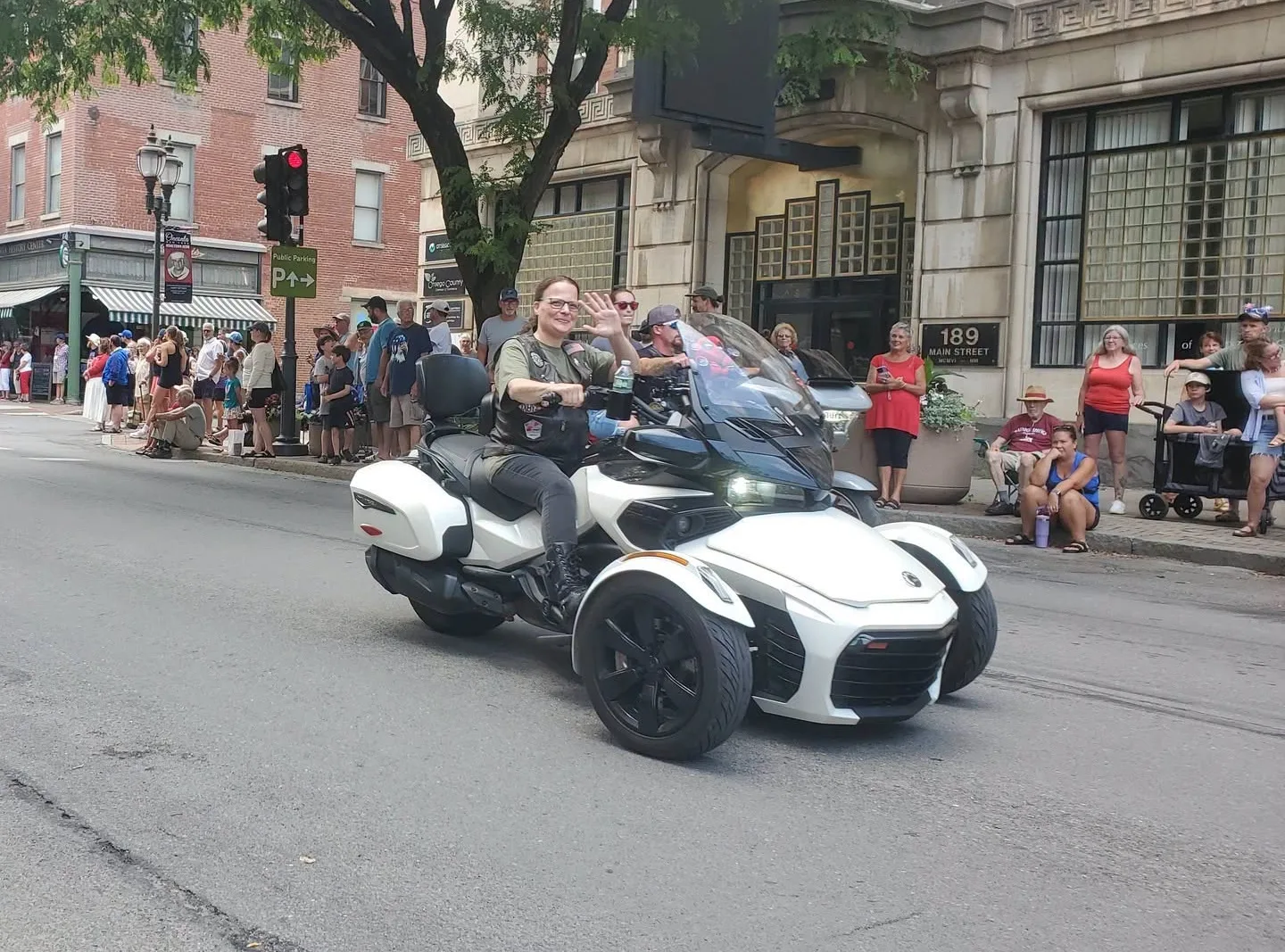 Angela Apostolakis waving from Selina, her white Can-Am Spyder F3T, as she rides a parade route past a crowd on Main Street