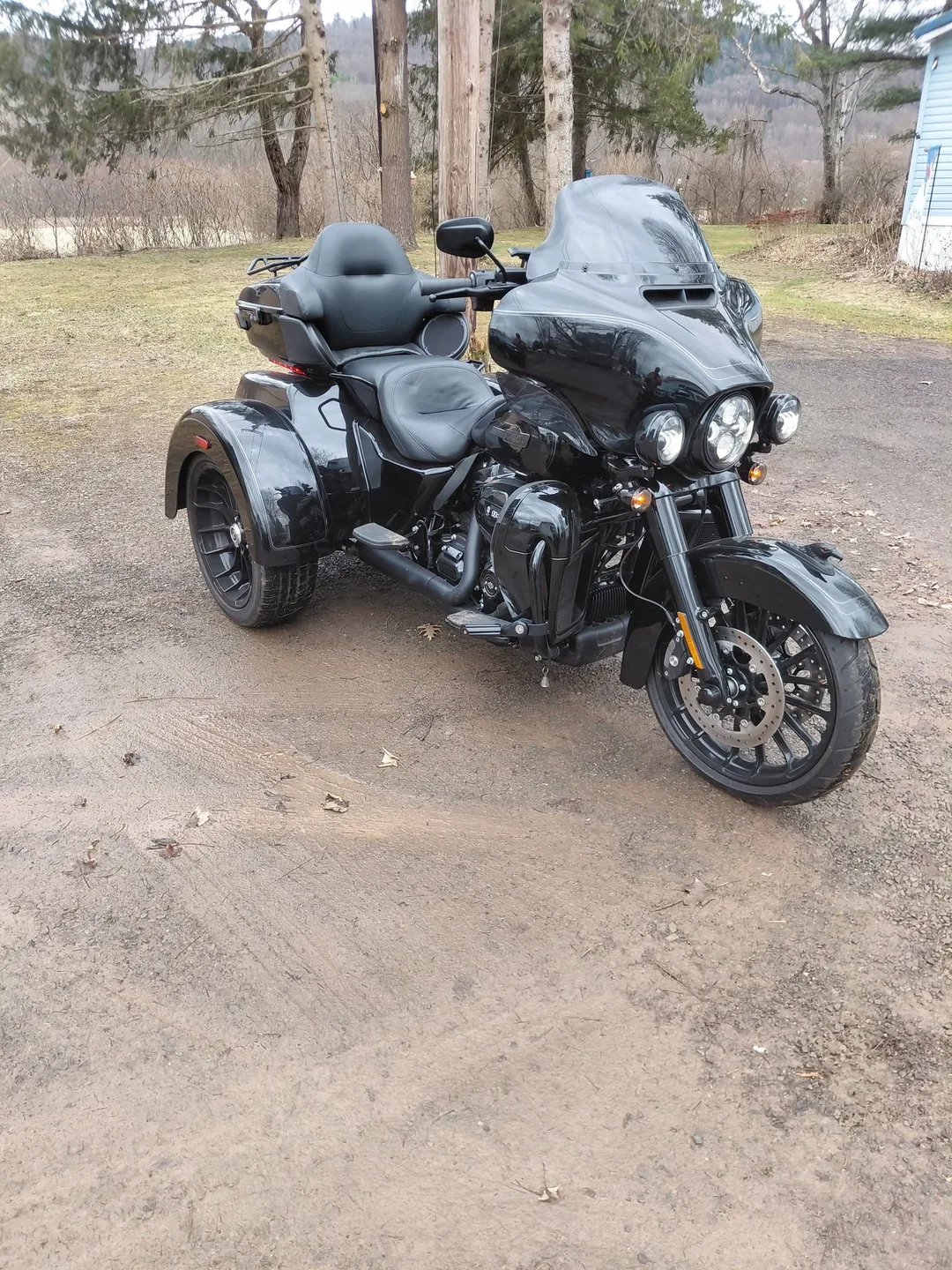 A blacked out Harley-Davidson trike with a batwing fairing, a passenger backrest and a luggage rack, parked on a gravel drive