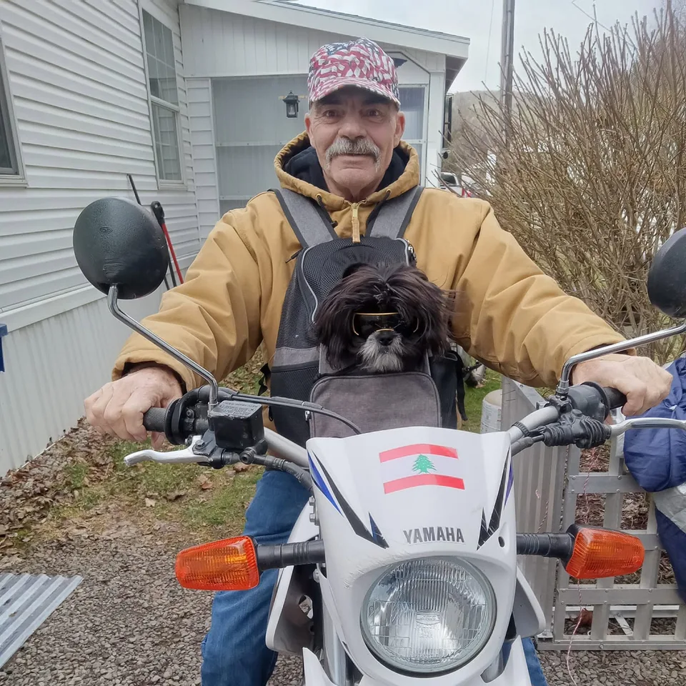 Arlo in goggles, tucked into a carrier worn on the chest of a rider in a tan jacket and a stars and stripes cap, sitting on a white Yamaha