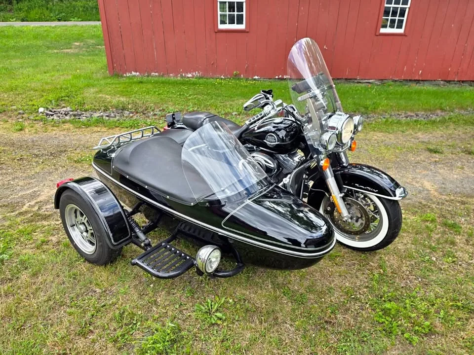 A black Harley-Davidson with a matching black sidecar, both behind clear windscreens, parked on the grass beside a red barn