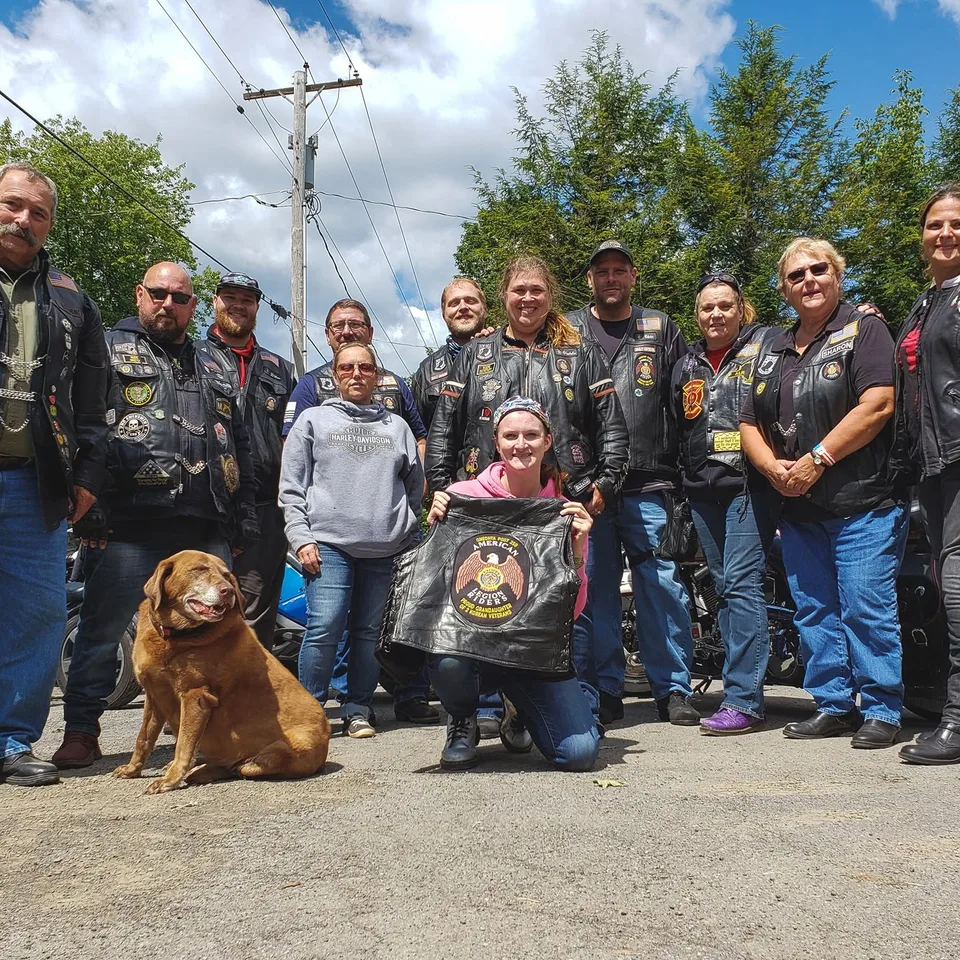 A dozen riders in their vests standing in front of their bikes on a summer day, with Charlie sitting in the middle of the group