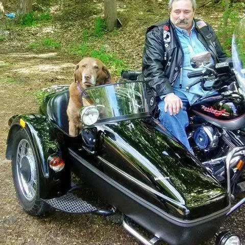 Charlie sitting in the sidecar of a black Harley-Davidson on a wooded drive, with a rider in a leather jacket alongside him