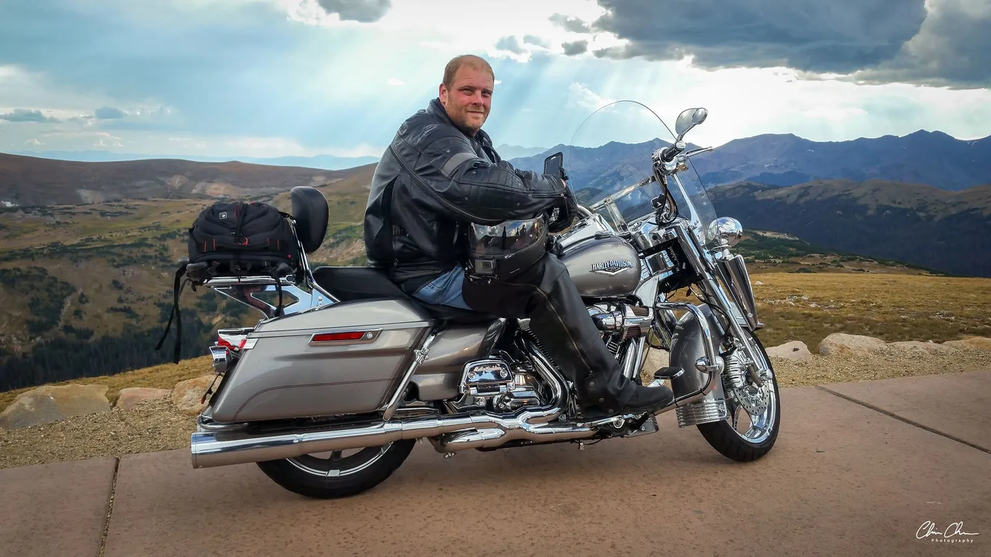 Chris Chase sitting on his silver Harley-Davidson Road King on a mountain overlook, with peaks and storm clouds behind him