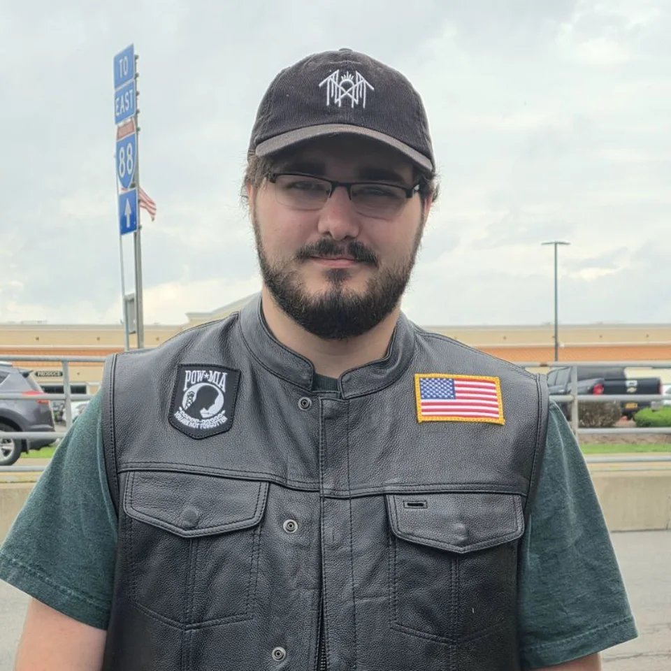 Connor Ouimet in his leather vest with POW/MIA and American flag patches, standing outdoors by Interstate 88