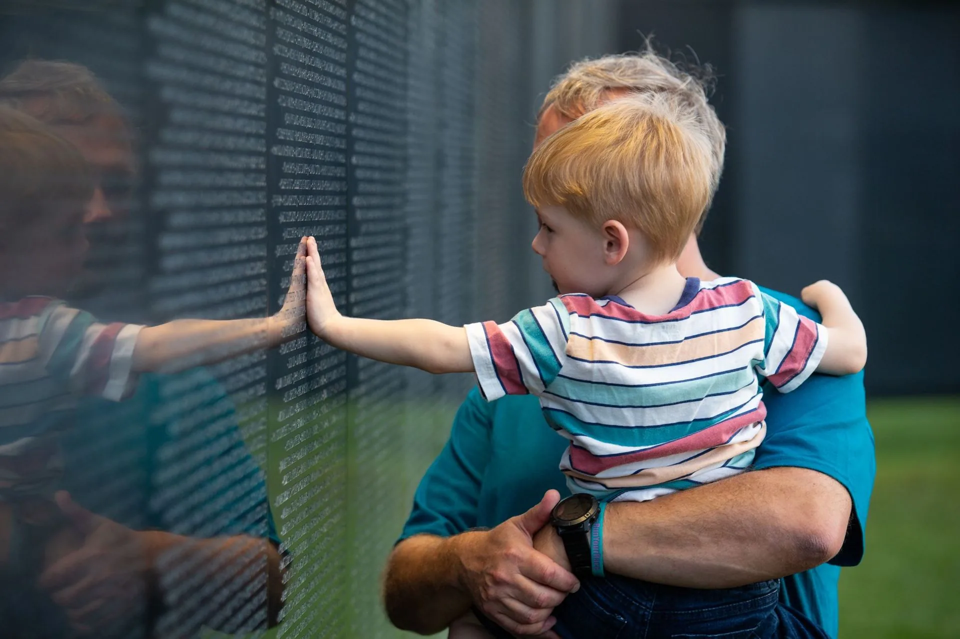A father holds his young son as the boy presses his hand to the names on The Wall That Heals