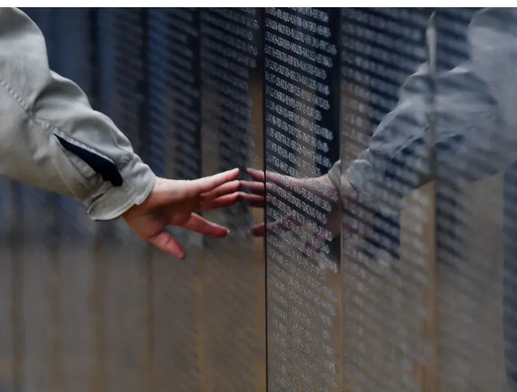 A visitor's hand rests on the names, reflected in the polished panels of The Wall
