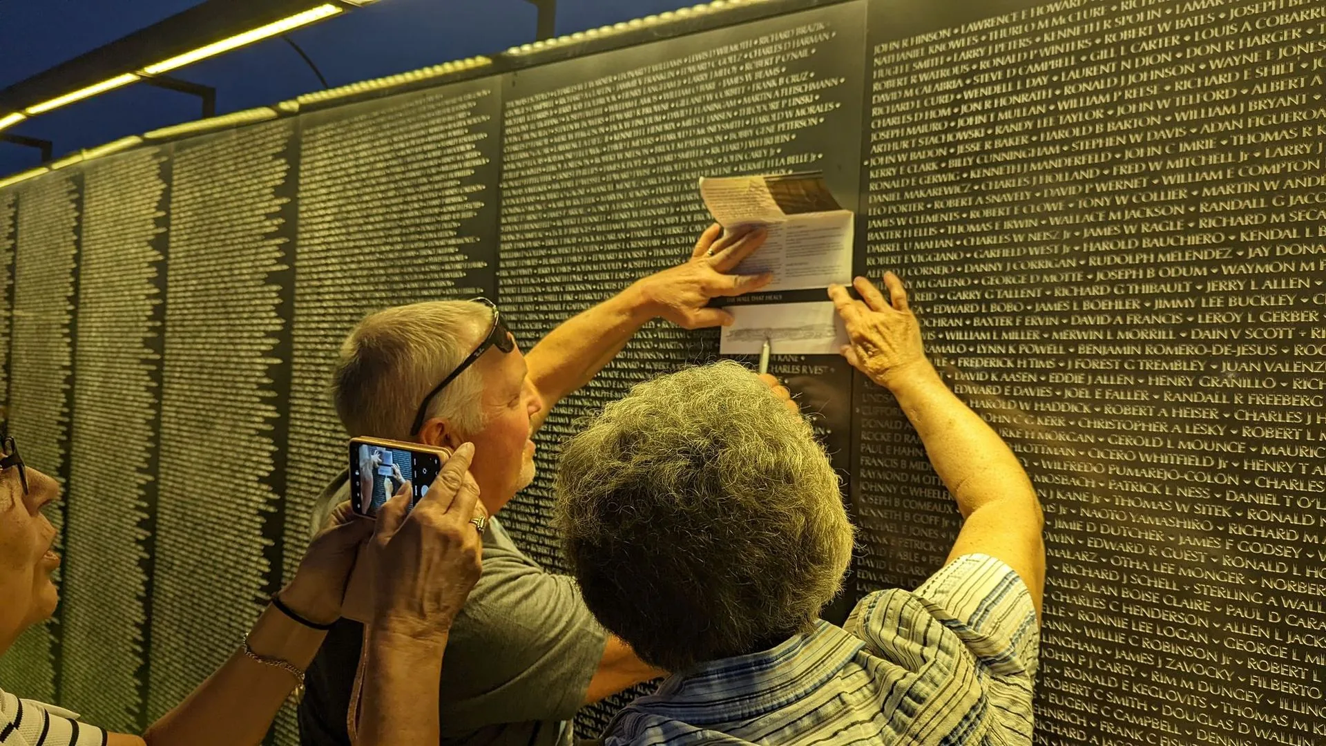 Family members make a name rubbing at night beneath the amber lights of The Wall